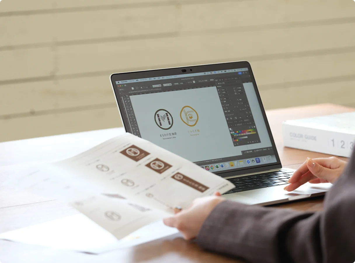ノートパソコンの画面でロゴデザイン制作をしている様子と資料を手に取るビジネスパーソンの手元